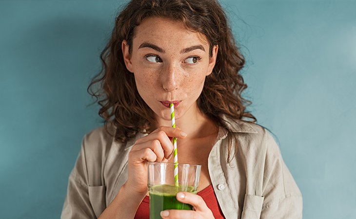 Young woman with freckles drinking a green drink through a straw in front of a blue background